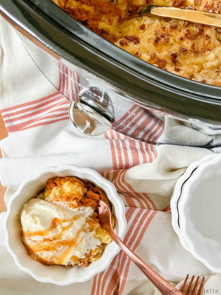 A bowl of dessert topped with whipped cream and caramel sauce sits on a striped cloth, beside a slow cooker filled with the same Thanksgiving treat. A fork rests in the bowl, ready to enjoy this festive delight.
