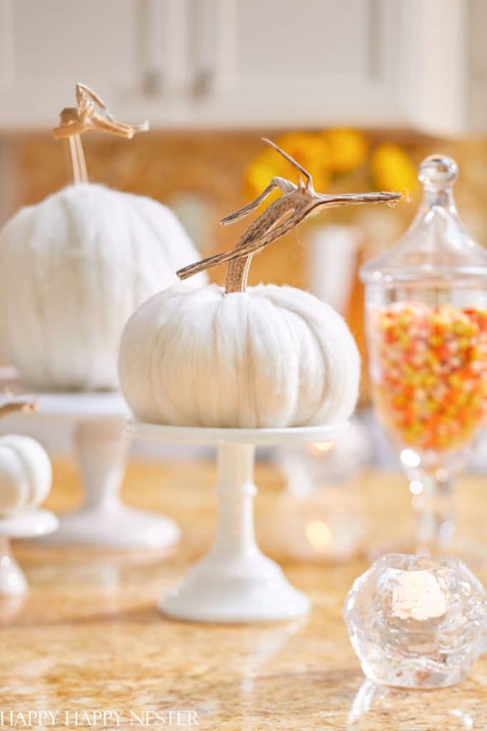 White velvet pumpkins with natural stems, perfect for white pumpkin crafts, are displayed on white cake stands beside a glass jar of candy corn and a small candle holder, set on a light-colored kitchen counter.