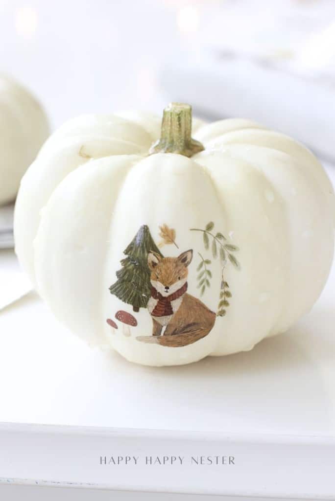 A white pumpkin decorated with a fox in a scarf, sitting by a pine tree and mushrooms—one of the cutest pumpkin decorating ideas without carving, using temporary tattoos for easy, mess-free charm.