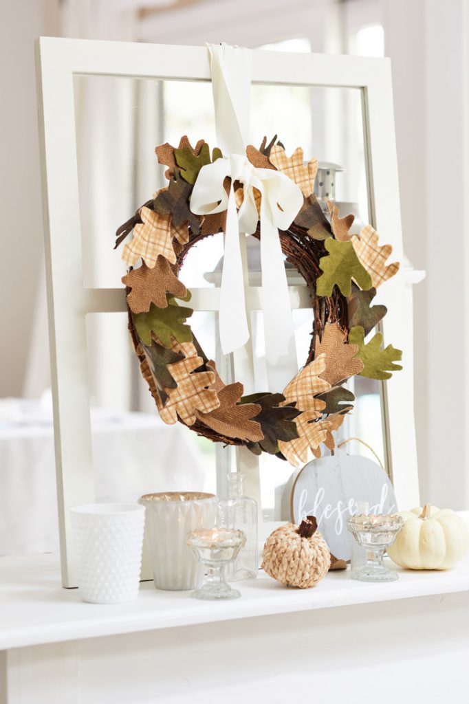 A decorative fall wreath—one of the loveliest wall crafts for fall—hangs on a white-framed mirror. In front are candles, a small "blessed" sign, a white pumpkin, and a textured ornament, all arranged on a white surface.