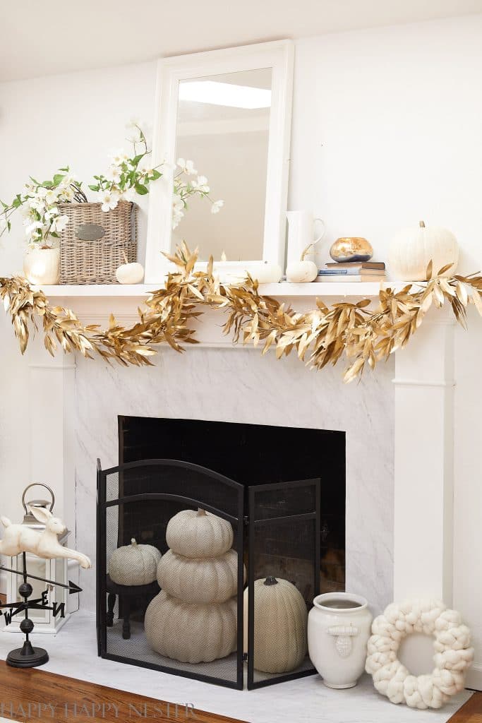A white fireplace mantel decorated with gold leaf garland, white pumpkins, a wicker basket with flowers, books, and a white-framed mirror. Cozy wall crafts for fall and stacked pumpkins add charm to the neutral-toned hearth decor.