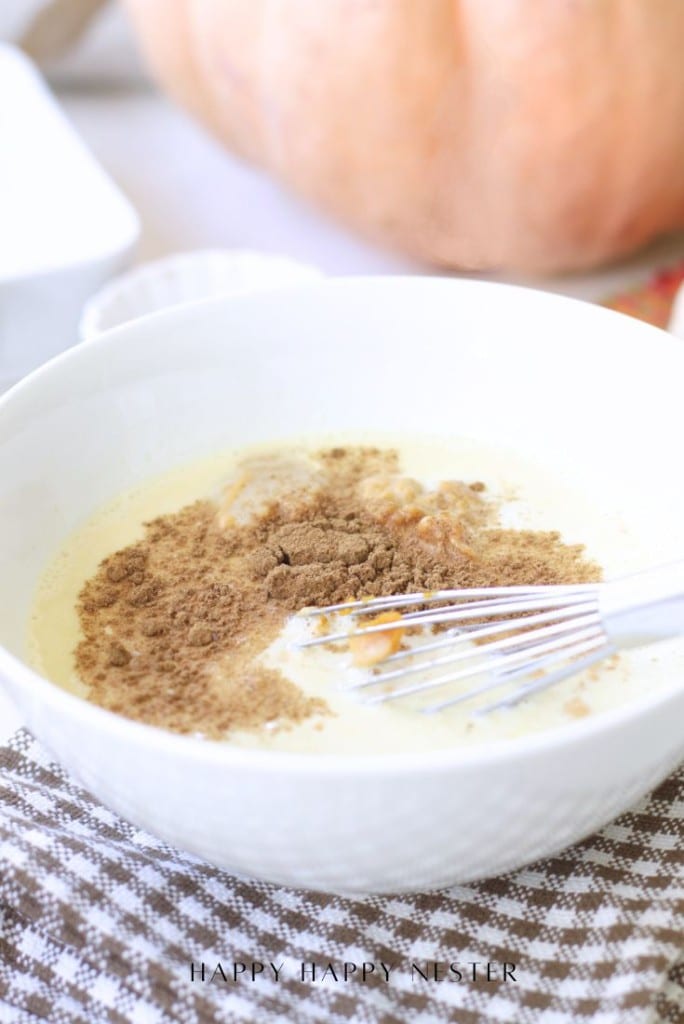 A white bowl with a creamy liquid mixture, brown spices, and a metal whisk on top, placed on a brown checkered cloth. A blurred pumpkin is in the background.