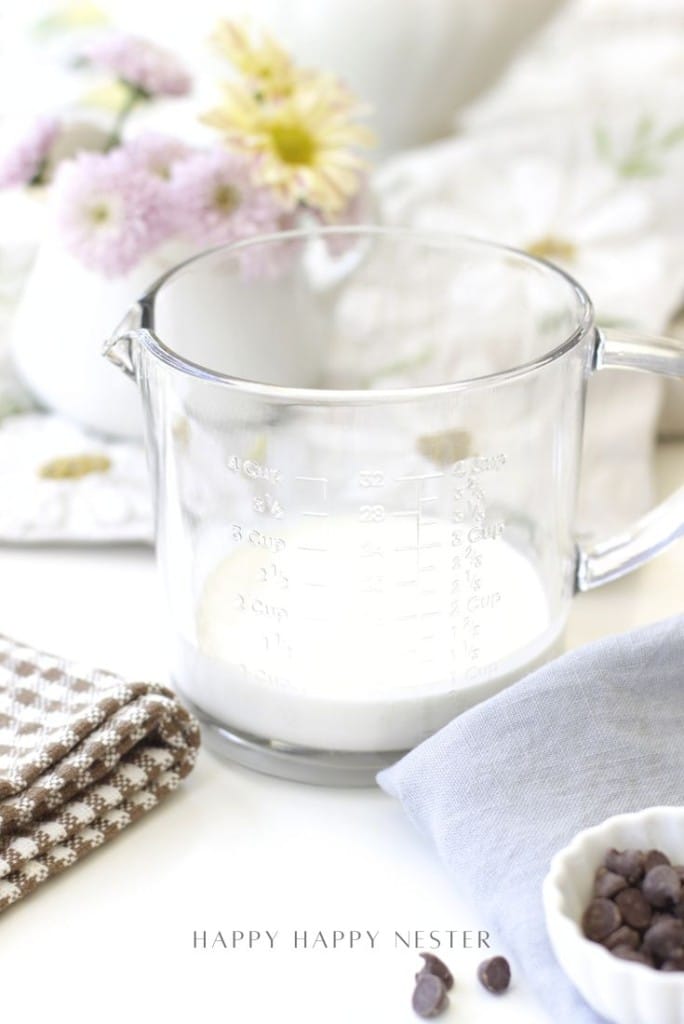 A glass measuring cup with milk sits on a white surface, surrounded by a brown checkered cloth, a gray napkin, a small bowl of chocolate chips—perfect for an easy chocolate ganache recipe—and a vase with light purple flowers.