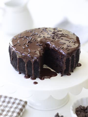 A round chocolate cake with glossy chocolate ganache and chocolate sprinkles sits on a white cake stand. A slice has been removed, revealing the moist interior. A checked cloth and small bowl of chocolate chips are nearby.