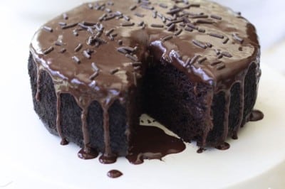 A round chocolate cake with glossy chocolate ganache and chocolate sprinkles sits on a white cake stand. A slice has been removed, revealing the moist interior. A checked cloth and small bowl of chocolate chips are nearby.