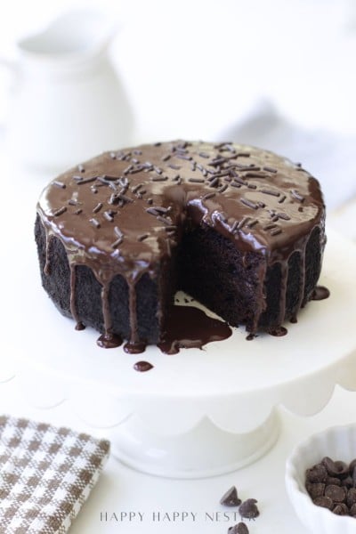 A round chocolate cake with glossy chocolate ganache and chocolate sprinkles sits on a white cake stand. A slice has been removed, revealing the moist interior. A checked cloth and small bowl of chocolate chips are nearby.