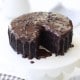 A round chocolate cake with glossy chocolate ganache and chocolate sprinkles sits on a white cake stand. A slice has been removed, revealing the moist interior. A checked cloth and small bowl of chocolate chips are nearby.