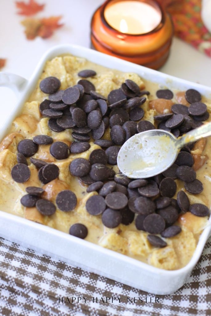A white dish filled with pumpkin bread pudding topped with dark chocolate chips. A spoon rests on top. In the background, a lit candle and autumn-themed decor complete this cozy pumpkin bread pudding recipe scene.