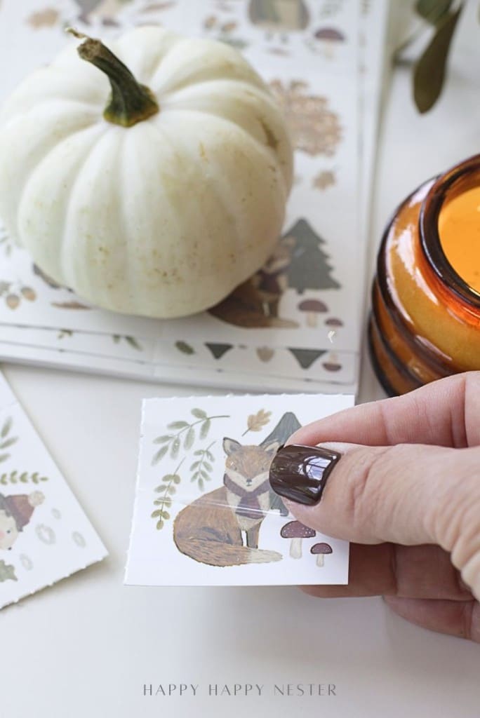 A hand with dark nail polish holds a small card featuring an illustrated fox, mushrooms, and leaves. Nearby are a white pumpkin, an amber candle, and more cards with autumn-themed designs.