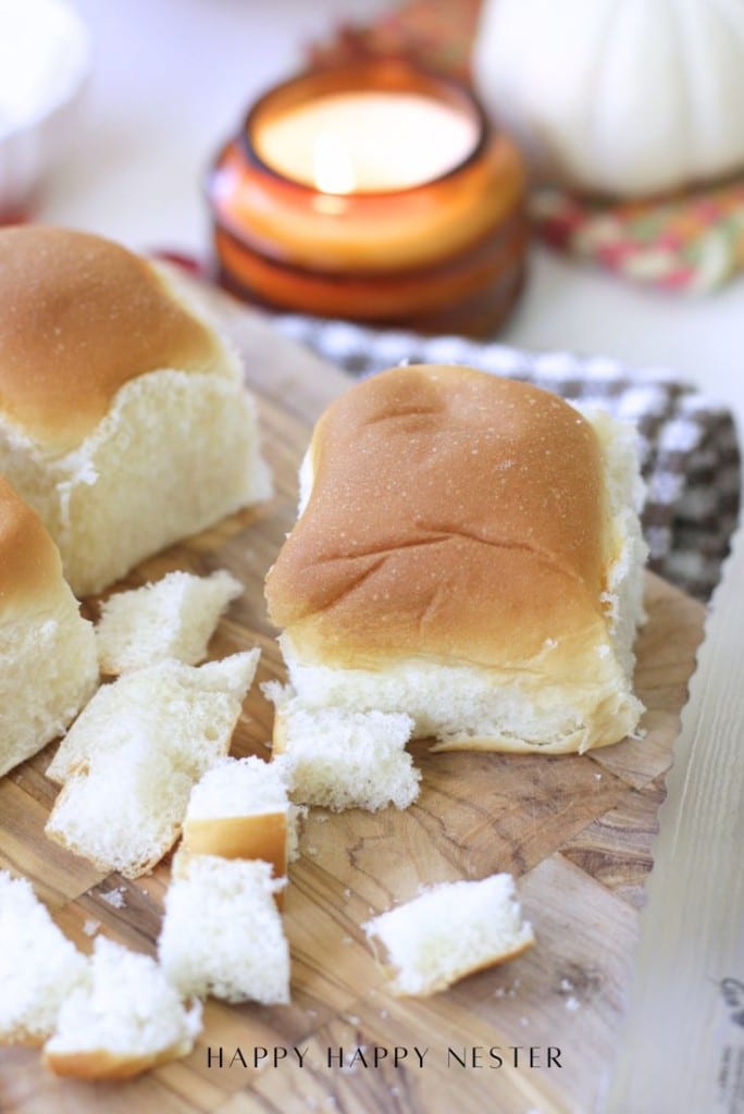 A soft dinner roll sits on a wooden cutting board beside small bread cubes—perfect for making a pumpkin bread pudding recipe. A lit candle and a white pumpkin are blurred in the background, adding to the cozy atmosphere.