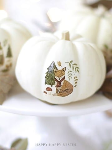 Three white pumpkins with delicate animal illustrations, created using temporary tattoos, sit on a white cake stand surrounded by dried leaves—a perfect example of pumpkin decorating ideas without carving in a soft, light-filled setting.