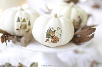 Three white pumpkins with delicate animal illustrations, created using temporary tattoos, sit on a white cake stand surrounded by dried leaves—a perfect example of pumpkin decorating ideas without carving in a soft, light-filled setting.