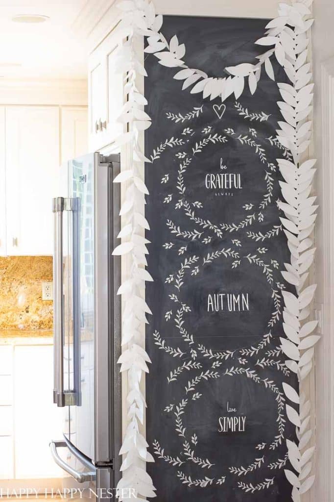 A chalkboard wall in a kitchen, decorated with white chalk wreaths and handwritten messages—“be GRATEFUL,” “AUTUMN,” and “live SIMPLY”—features wall crafts for fall, including white paper leaf garlands draped around the board.