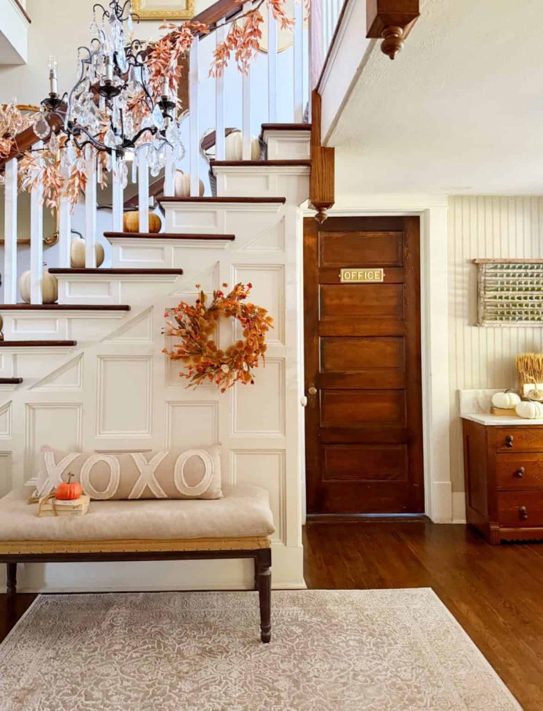 A cozy entryway with a wooden staircase decorated with autumn leaves, pumpkins, wall crafts for fall, and an orange wreath. A bench with "XOXO" pillows sits on a patterned rug near a wooden "OFFICE" door.