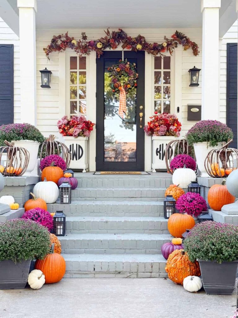 A front porch decorated for fall with orange, white, and purple pumpkins, potted purple and pink flowers, lanterns, and touches of white pumpkin crafts, plus a garland and wreath with autumn leaves and flowers on the door and railing.