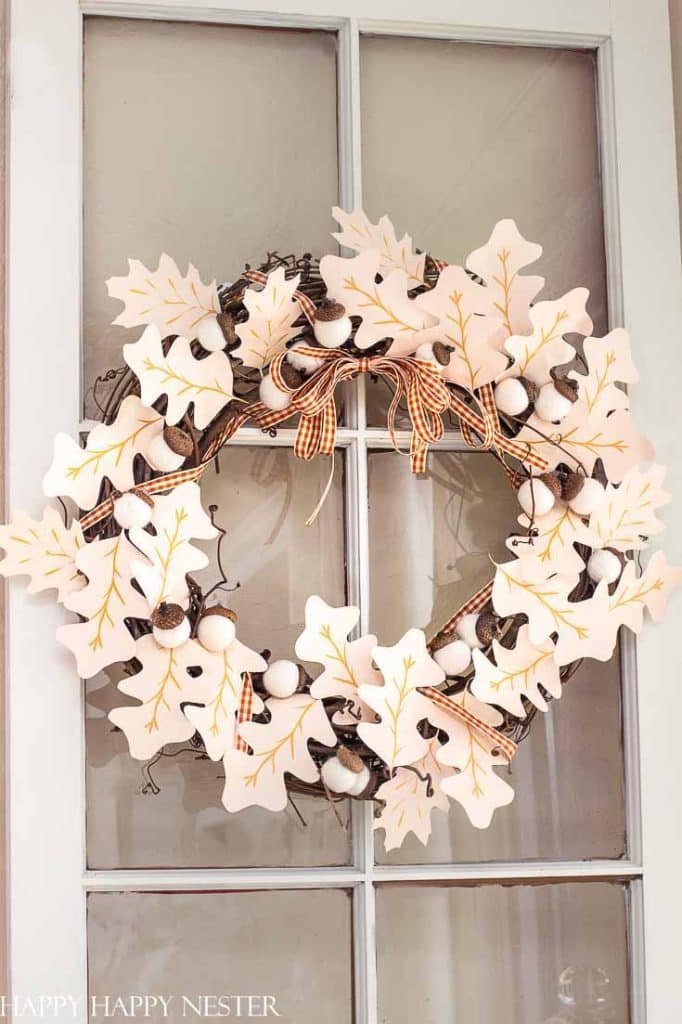 A wreath made of white and beige paper oak leaves, brown acorns, and white pom-poms hangs on a glass-paneled door, decorated with small orange plaid ribbon bows—my favorite Thanksgiving decor for a festive touch.