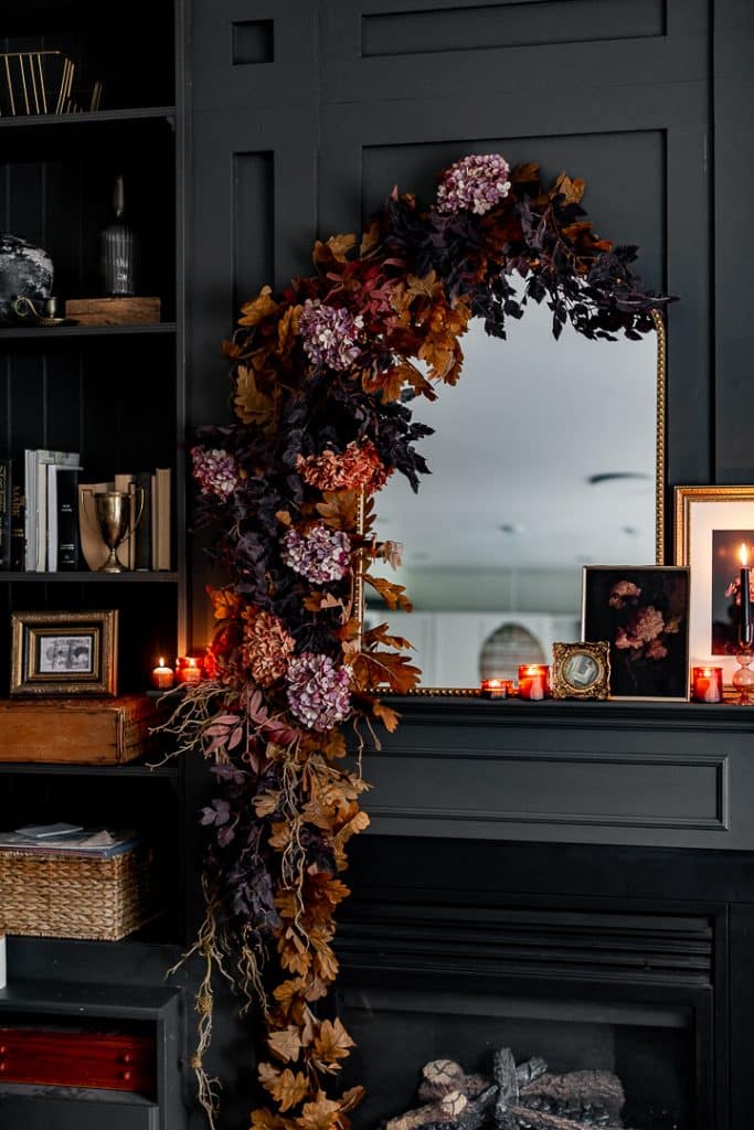 A dark mantel decorated with a fall floral garland of purple and brown leaves and flowers—like wall crafts for fall—draped over a mirror, surrounded by books, framed photos, and red candles, creating a cozy, moody atmosphere.