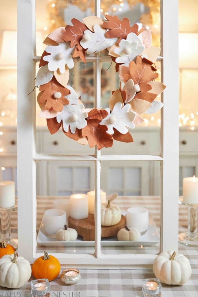 A fall wreath made of felt leaves in white, copper, and brown hangs on a window frame. Small pumpkins, candles, and my favorite Thanksgiving decor are arranged on a checkered table beneath the wreath.