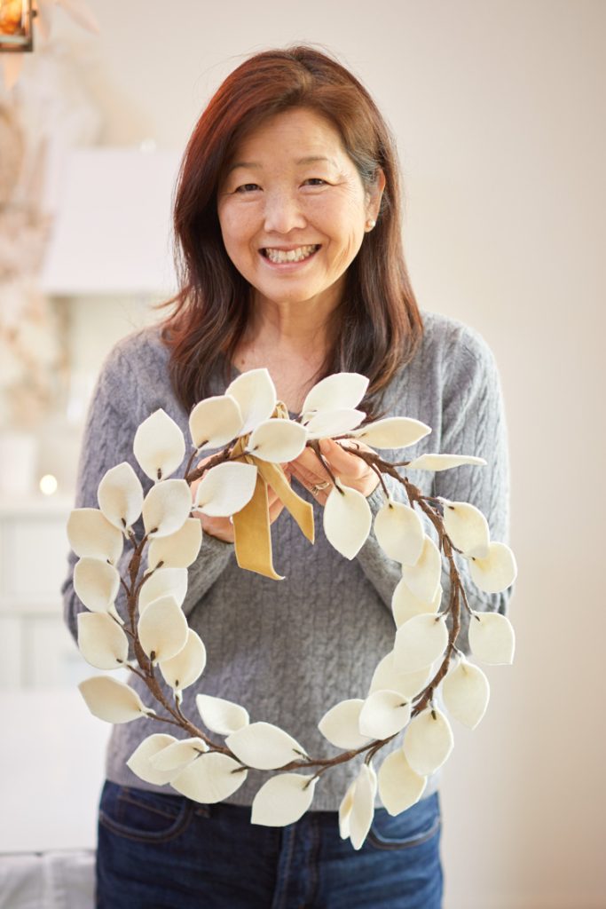 A smiling woman with long brown hair, wearing a gray sweater, holds a handmade wreath decorated with white felt leaves and a small gold ribbon—perfect wall crafts for fall. The background is softly blurred.