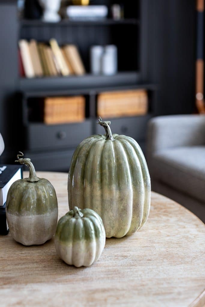 Three decorative pumpkins in muted green and cream tones showcase creative pumpkin decorating ideas on a wooden table, with a blurred bookshelf and armchair in the background, creating a cozy, autumn-inspired atmosphere.