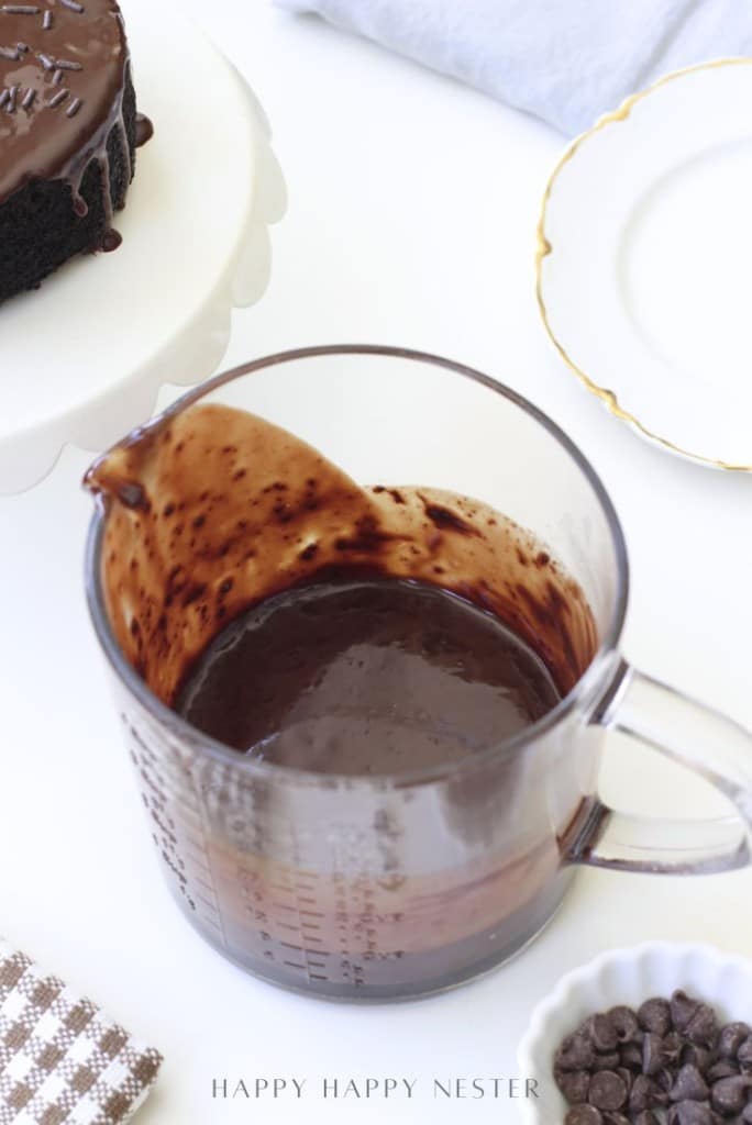 A glass measuring cup filled with freshly made chocolate ganache sits on a white surface near a cake on a stand, a plate, and a bowl of chocolate chips.
