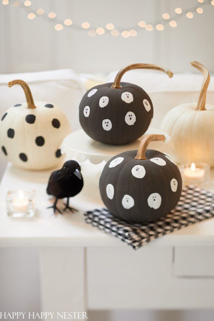 Black and white pumpkin crafts decorated with painted polka dots and ghost faces sit on a white table with candles, a small black bird figurine, and string lights in the background.