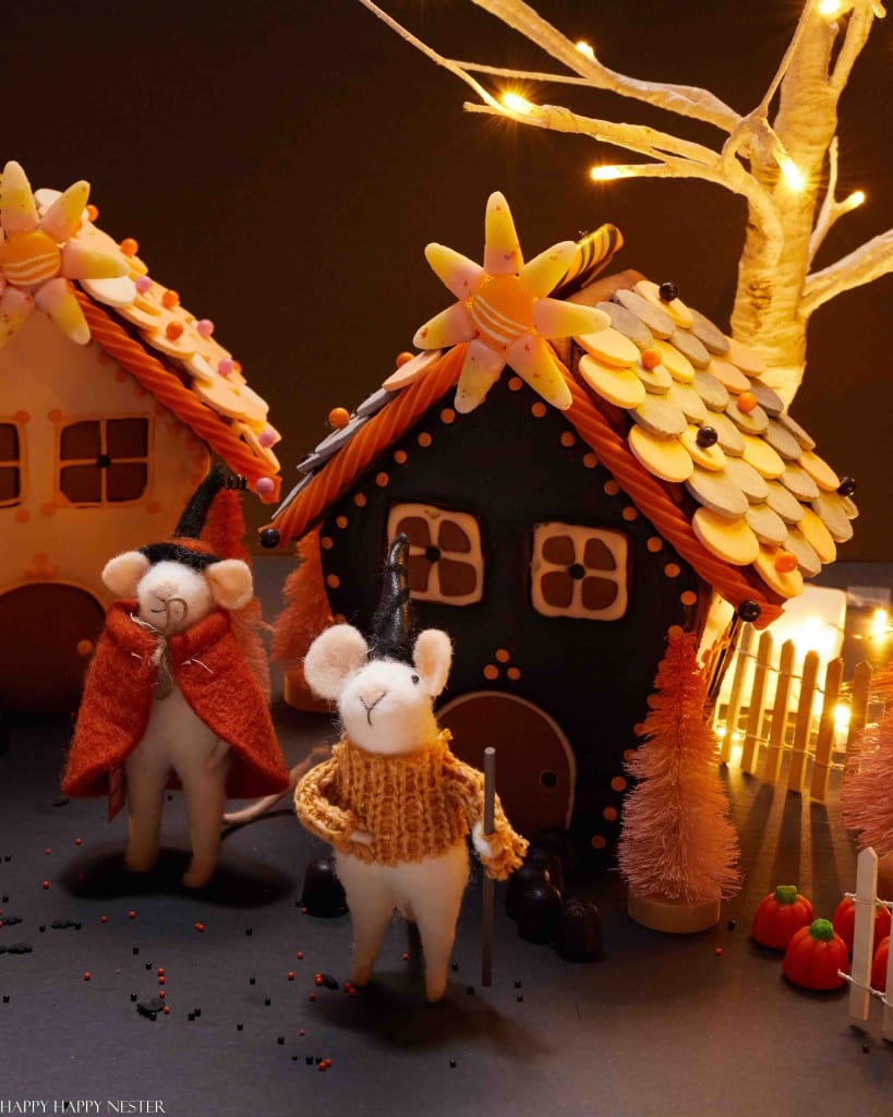 Two felt mice in cozy sweaters and witch hats stand before whimsical gingerbread houses decorated with buttons, candy roofs, and halloween treats. Warm fairy lights and small festive pumpkins complete the enchanting scene.