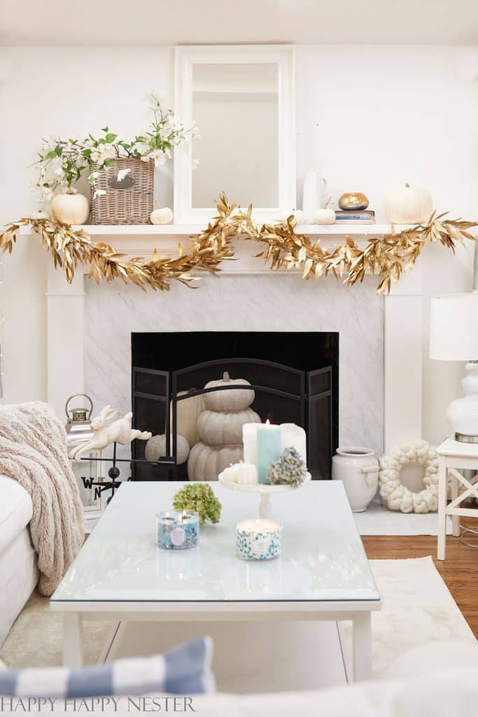 A bright living room with a white marble fireplace decorated in my favorite Thanksgiving decor—gold garland, white pumpkins, and soft fall accents. A glass coffee table holds candles, creating a cozy, elegant atmosphere.