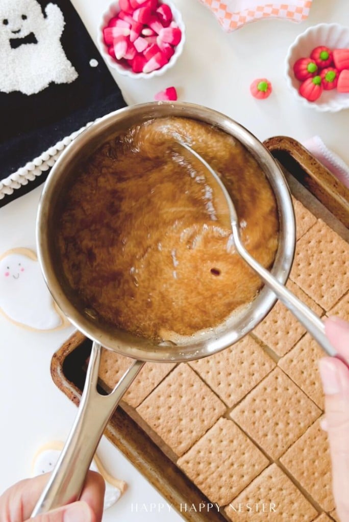 A person pours a bubbly, caramel-like mixture from a saucepan over a tray of graham crackers. Decorative bowls with candies and ghost-shaped cookies are nearby, suggesting a festive baking scene.
