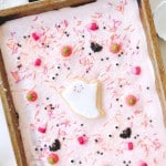 A rectangular sheet cake with pink frosting is decorated with Halloween-themed sprinkles, candy eyes, and a central ghost-shaped cookie—perfect for pairing with your favorite Halloween bark recipe (easy). The cake sits in a metal pan on a festive table.