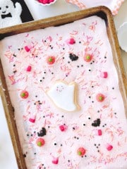 A rectangular sheet cake with pink frosting is decorated with Halloween-themed sprinkles, candy eyes, and a central ghost-shaped cookie—perfect for pairing with your favorite Halloween bark recipe (easy). The cake sits in a metal pan on a festive table.