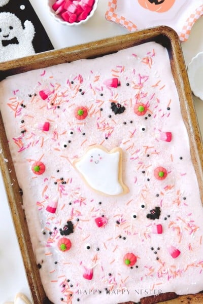 A rectangular sheet cake with pink frosting is decorated with Halloween-themed sprinkles, candy eyes, and a central ghost-shaped cookie—perfect for pairing with your favorite Halloween bark recipe (easy). The cake sits in a metal pan on a festive table.