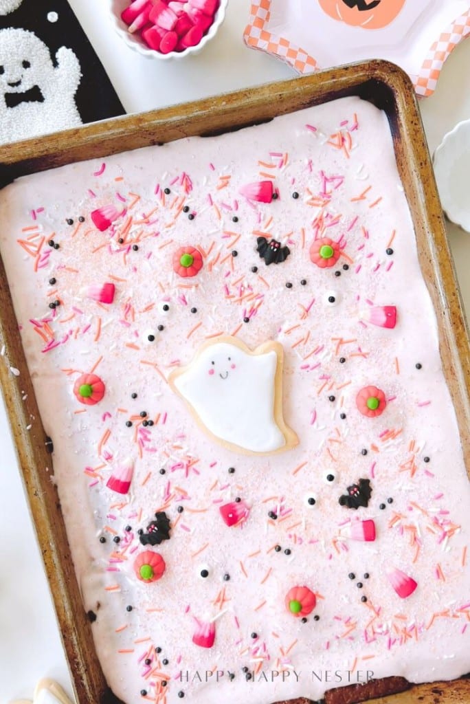 A rectangular sheet cake with pink frosting is decorated with Halloween-themed sprinkles, candy eyes, and a central ghost-shaped cookie—perfect for pairing with your favorite Halloween bark recipe (easy). The cake sits in a metal pan on a festive table.