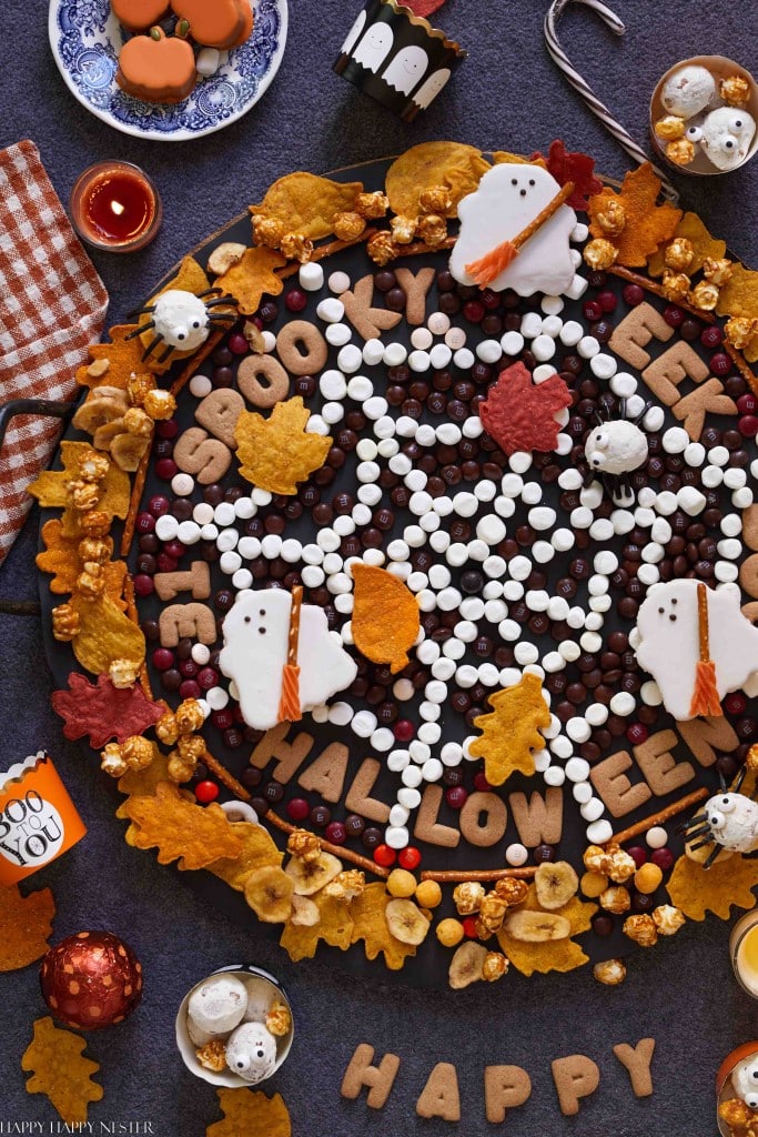 A Halloween-themed dessert platter features ghost-shaped cookies, halloween treats, and cookies spelling "SPOOKY," "EEK," and "HALLOWEEN" on a spiderweb of white candies, with autumn leaves and festive treats all around.