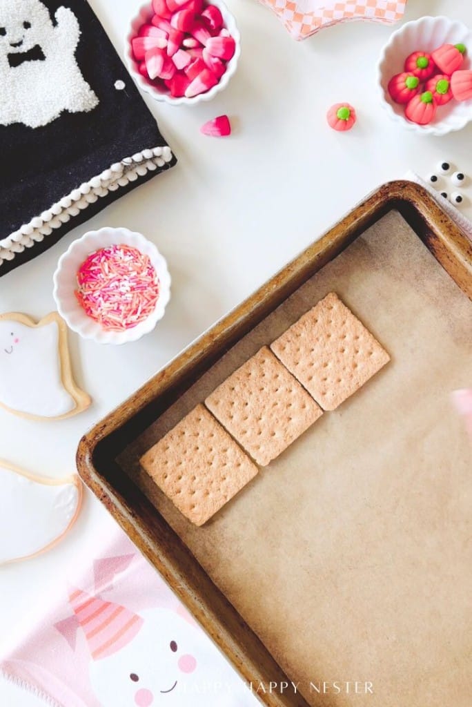 Three graham crackers are lined up in a baking pan with parchment paper. Surrounding the pan are bowls of pink sprinkles, candy, a ghost cookie, and black and white towels with playful Halloween themes.