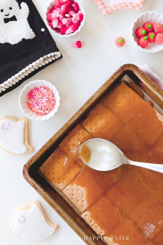A baking pan with graham crackers being topped with caramel using a spoon is on a white surface. Nearby are ghost-shaped cookies, pink sprinkles, Halloween candies, and a black towel with a white ghost design.