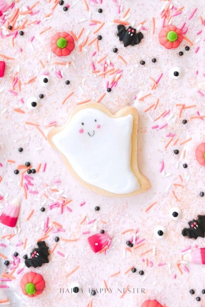 A ghost-shaped sugar cookie with white icing and a smiling face is surrounded by Halloween-themed sprinkles, candy bats, eyeballs, and candy corn—perfect inspiration for an easy Halloween bark recipe on a playful pink background.