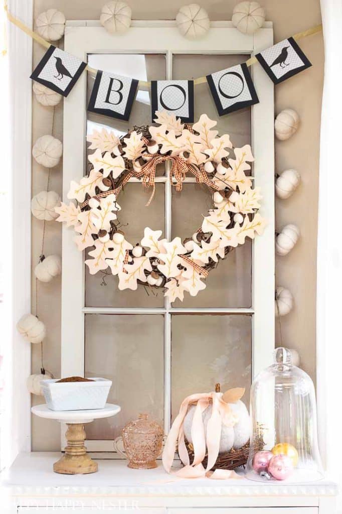 A decorative Halloween display features wall crafts for fall like a “BOO” banner, a wreath with leaves and berries on an old window, white pumpkins, a glass cloche with ornaments, a teapot, and a cake stand on a shelf.