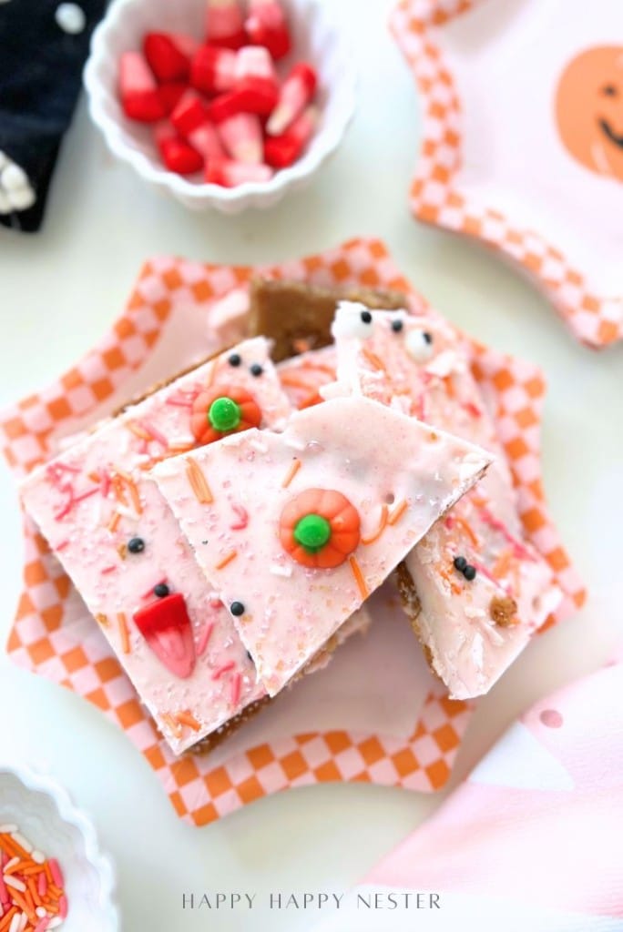 A stack of pink frosted cookie bars, inspired by an easy Halloween bark recipe, is decorated with candy eyes, sprinkles, and gummy worms on a checkered plate, surrounded by bowls of sprinkles and chopped strawberries.