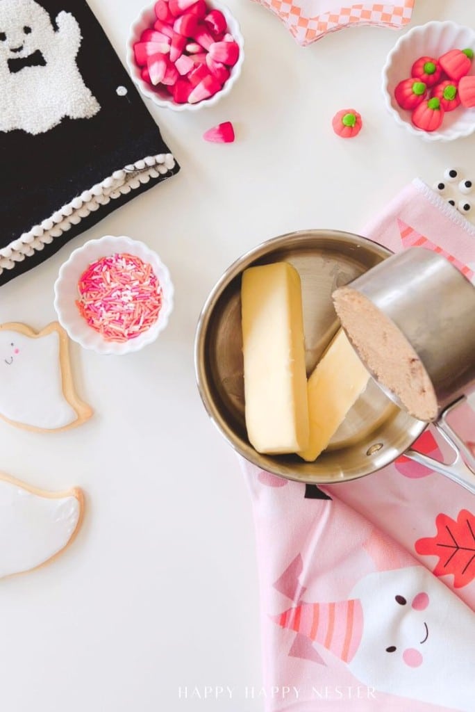 A saucepan with two sticks of butter sits on a pink towel. Nearby are Halloween cookies shaped like ghosts, bowls of sprinkles, candy, and a decorated black notebook—perfect inspiration for an easy Halloween bark recipe.