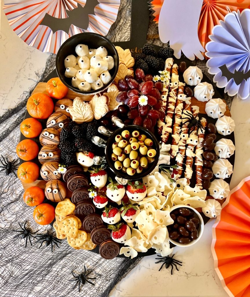 A Halloween-themed snack board featuring halloween treats like ghost meringues, chocolate-covered strawberries, stuffed olives, cookies, crackers, grapes, mini oranges with googly eyes, blackberries, and festive decorations.