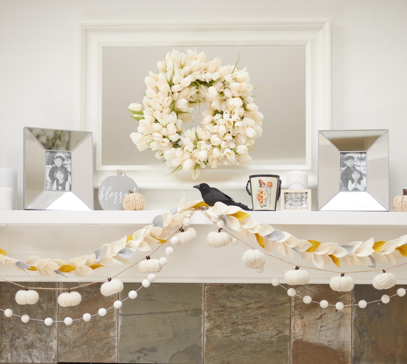 A white mantel decorated with a wreath of white tulips, framed photos, wall crafts for fall, small pumpkins, a black bird figurine, and garlands of white pumpkins and pom-poms creates a festive autumn display.