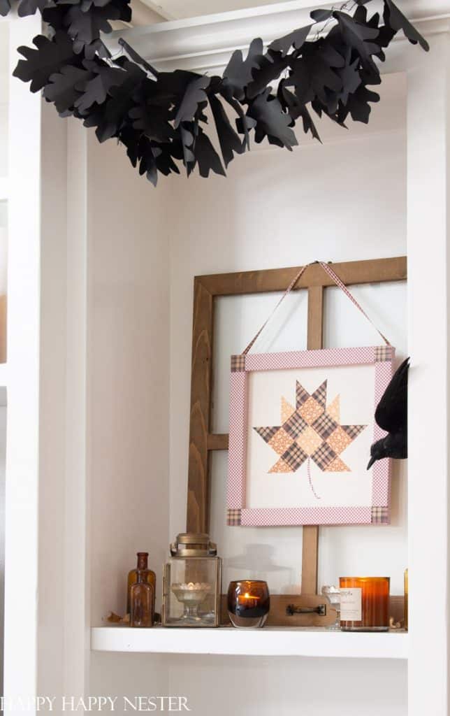 A cozy shelf decorated for fall with brown bottles, amber candles, a lantern, and framed patchwork leaf art—my favorite Thanksgiving decor. Above, a black leaf garland hangs across the white shelf.