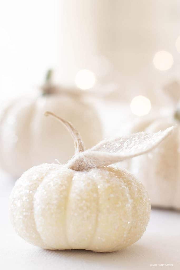 Small white pumpkin covered in sparkling glitter with two felt leaves, perfect for white pumpkin crafts, with more blurred white pumpkins softly glowing in the background.