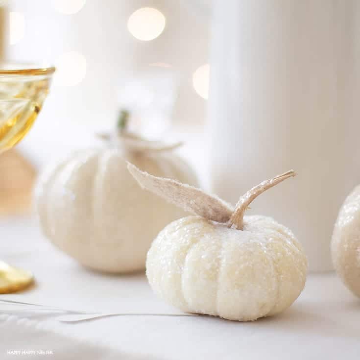 A small white pumpkin with a frosted, sparkling surface sits on a white tablecloth—my favorite Thanksgiving decor—surrounded by similar pumpkins and soft, out-of-focus lights in the background.