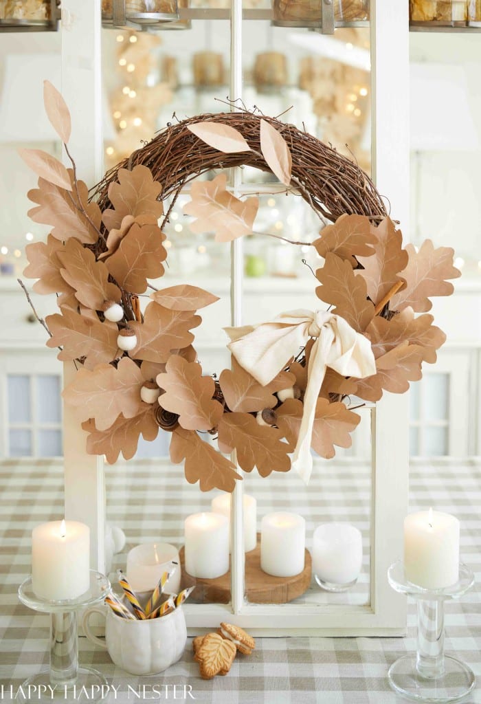 A rustic wreath made of brown paper oak leaves and white accents, ideal as wall crafts for fall, hangs on a glass window. It features a cream-colored bow, while white candles and autumn cookies rest on a checkered tablecloth below.