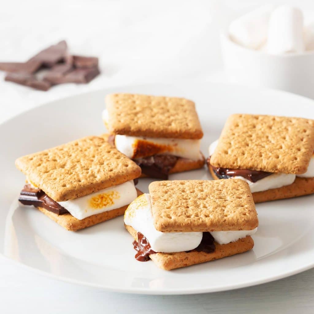 Four s’mores made with toasted marshmallows, melted chocolate, and graham crackers are arranged on a white plate—perfect as Halloween treats. In the background are pieces of chocolate and a bowl of marshmallows.