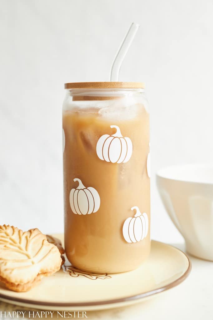 A glass of iced coffee with a bamboo lid and straw, decorated with white pumpkin illustrations—my favorite Thanksgiving decor—sits on a plate with a leaf-shaped cookie. A white bowl is in the background.