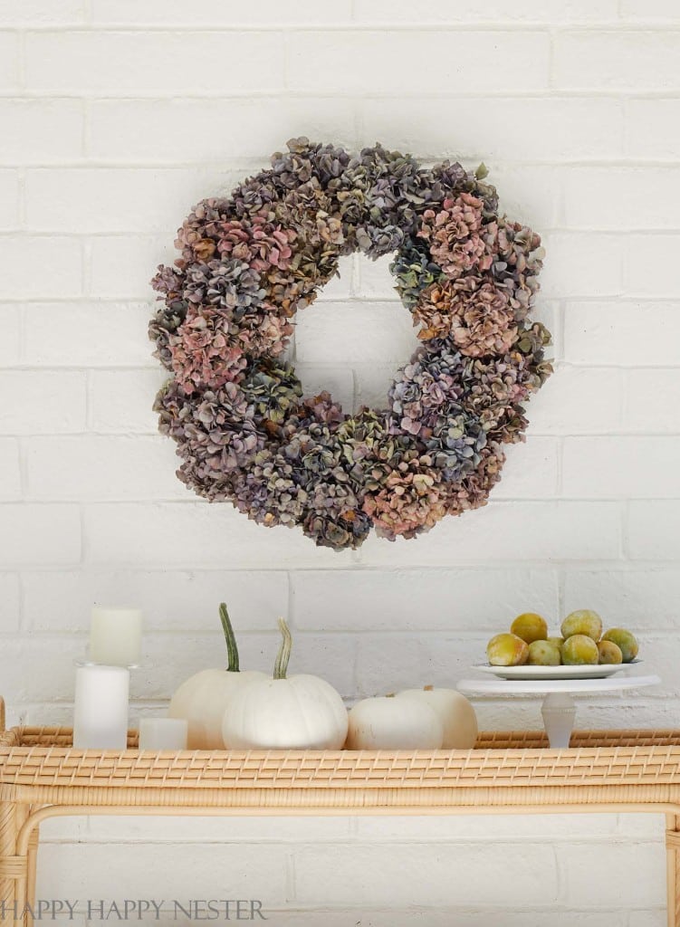 A dried hydrangea wreath, perfect for wall crafts for fall, hangs on a white brick wall above a wicker shelf with white pumpkins, candles, and a plate of yellow-green plums.