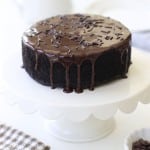 A round chocolate cake with glossy chocolate ganache made from an easy chocolate ganache recipe and chocolate sprinkles sits on a white cake stand. A patterned napkin and a small bowl of chocolate chips are nearby on a white surface.